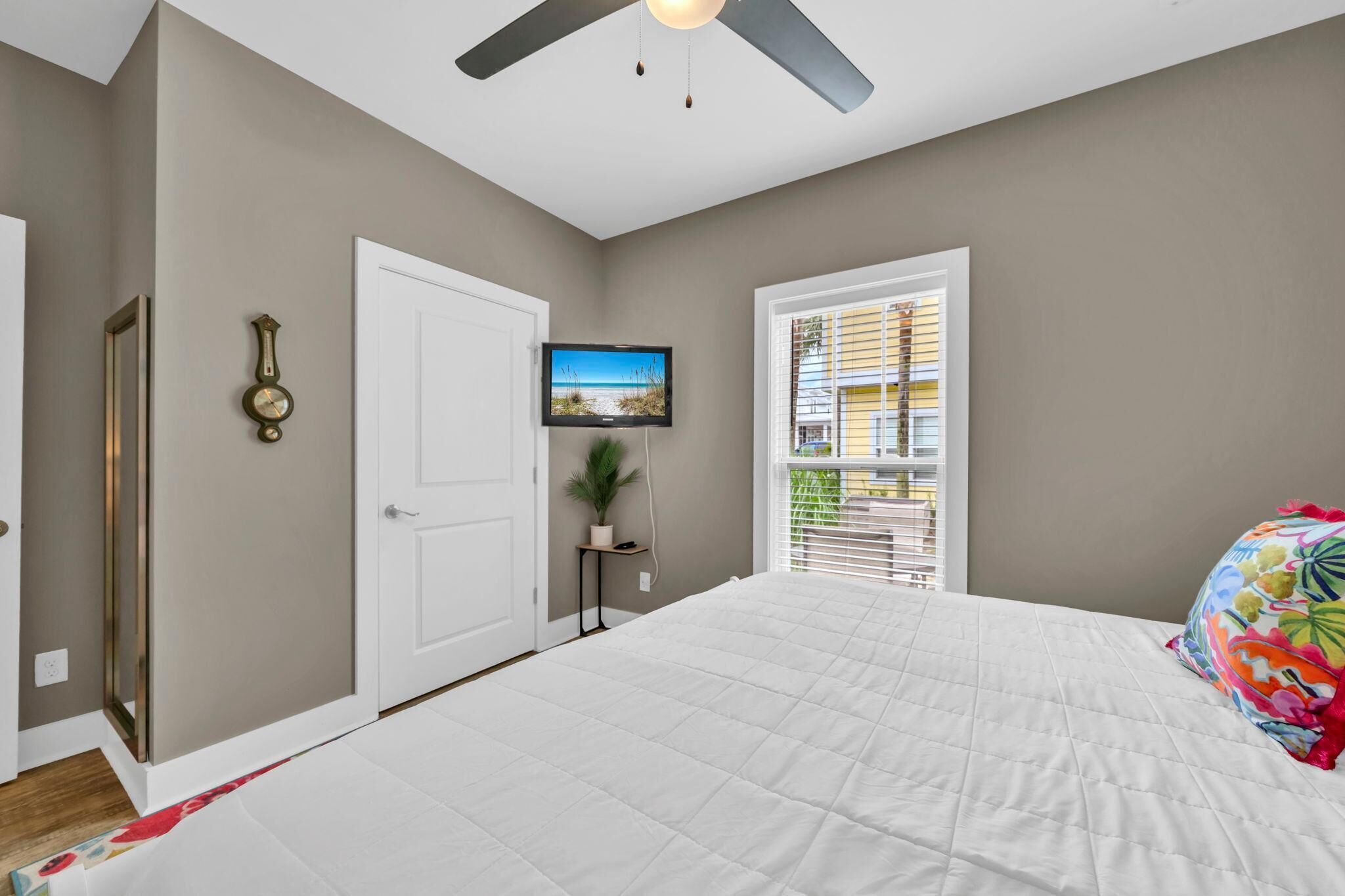 A bedroom with a bed, a ceiling fan, a TV, a door, and a window with blinds.