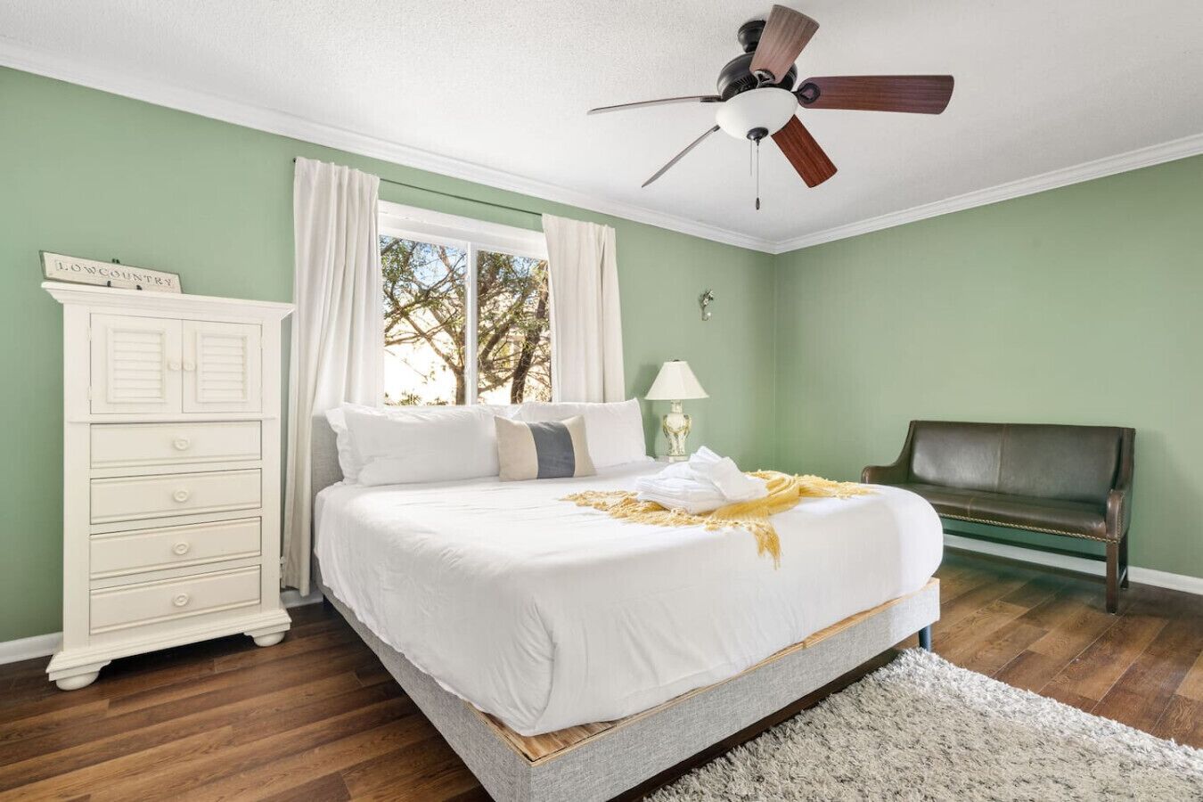 A bedroom with a bed, a dresser, a bench, a ceiling fan, and a window with curtains.