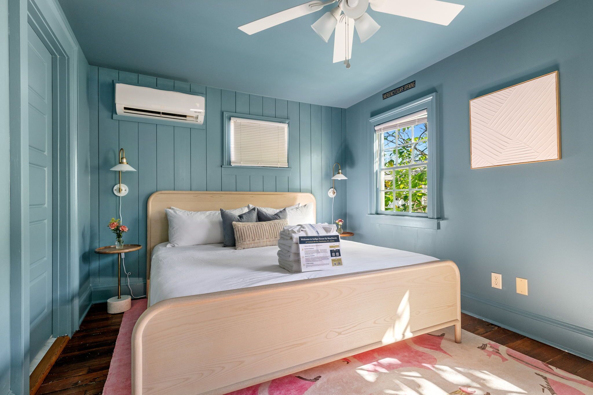 A bedroom with a bed, a ceiling fan, an air conditioning unit, a window with blinds, and a wall-mounted light fixture.
