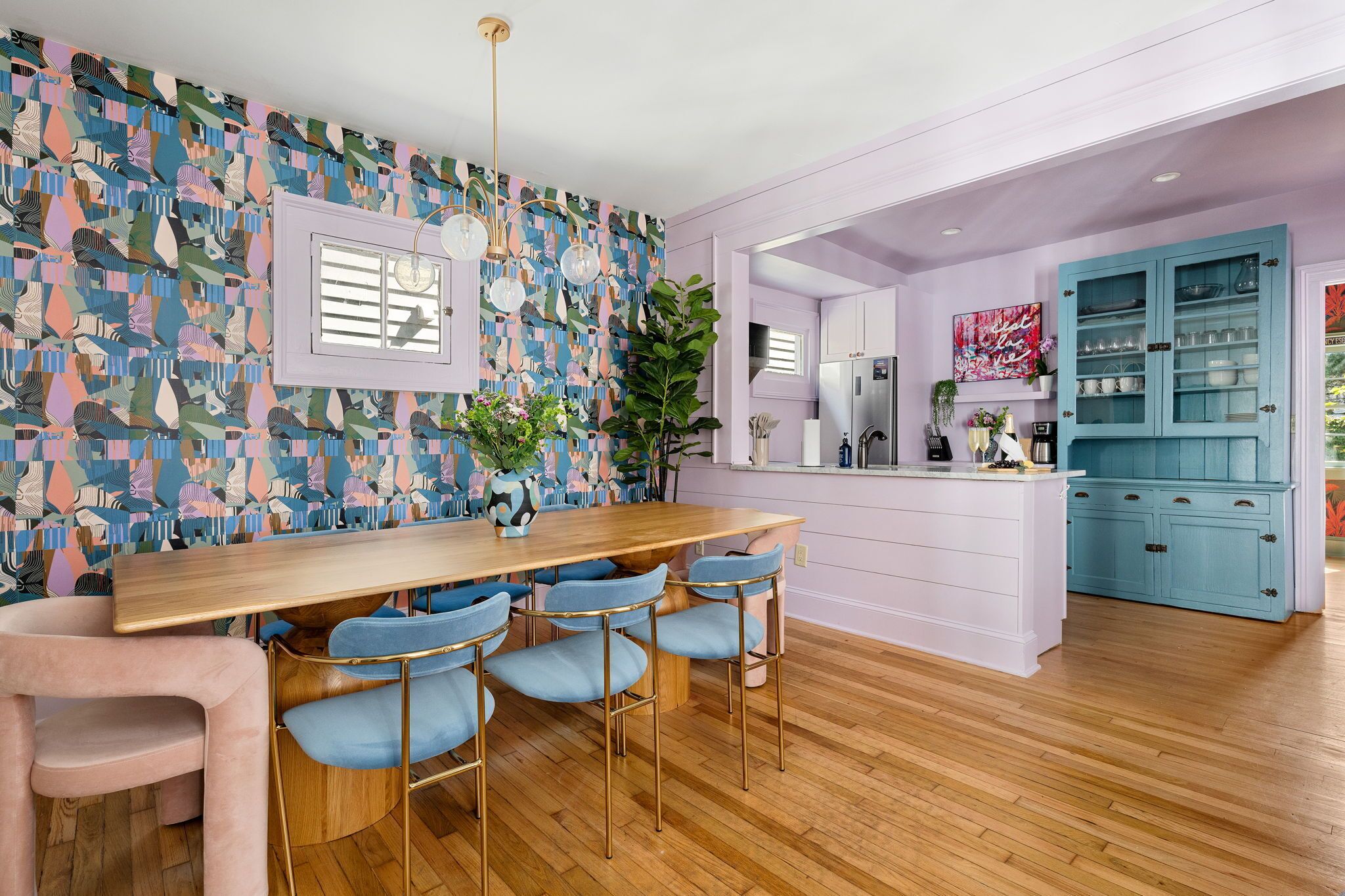 Une cuisine moderne avec une table à manger en bois, des chaises bleues et un mur décoré d’une œuvre abstraite aux couleurs vives.