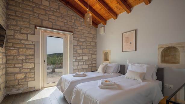 A bedroom with two beds, stone walls, a wooden ceiling, and a door leading to an outdoor area.