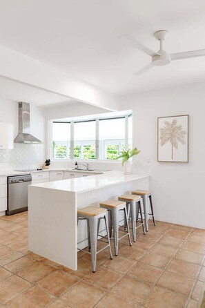 A modern kitchen with a central island, bar stools, a ceiling fan, and a window with a view of greenery. - Pura Vida Noosaville Retreat with Pool (Noosaville)