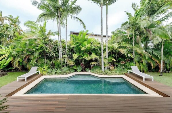 A rectangular swimming pool with two lounge chairs on wooden decking, surrounded by lush tropical vegetation and palm trees.