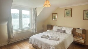 Un dormitorio con una cama grande, una ventana con cortinas, una mesita de noche con lámpara y cuadros en la pared.