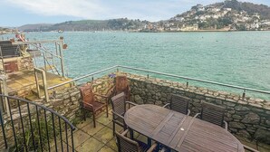 Una terraza con muros de piedra, muebles de madera y vistas a un mar tranquilo y colinas lejanas.