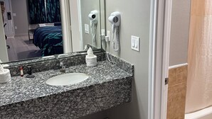 A bathroom with a granite countertop, a sink, two hair dryers mounted on the wall, and a trash can. - Scottish inns & suites (Houston)