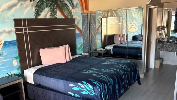 A modern hotel room with a large bed, a desk, and a chair. The room features a mural of a tropical beach scene on the ceiling. - Scottish inns & suites (Houston)