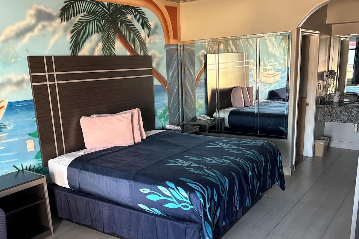 A modern hotel room with a large bed, a desk, and a chair. The room features a mural of a tropical beach scene on the ceiling.