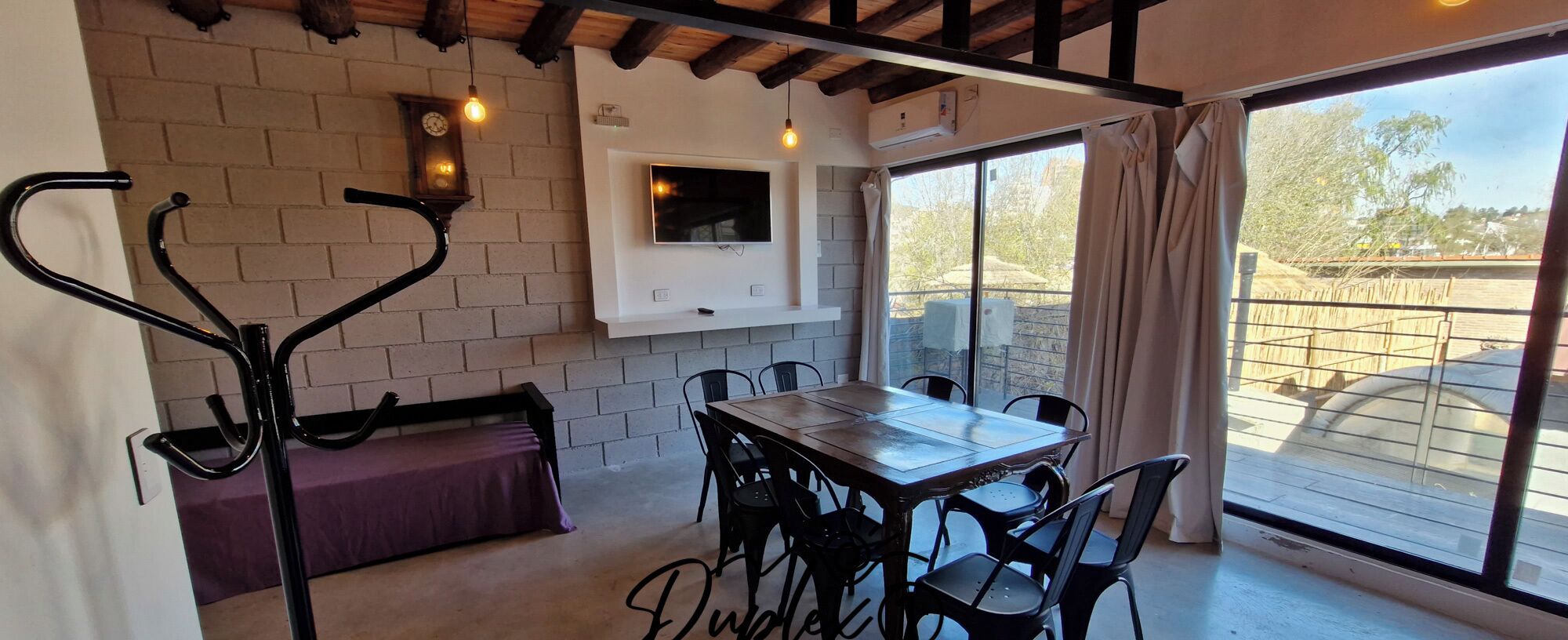 Zona pranzo con tavolo e sedie in legno, una panca, televisore a parete e un'ampia finestra con tende.