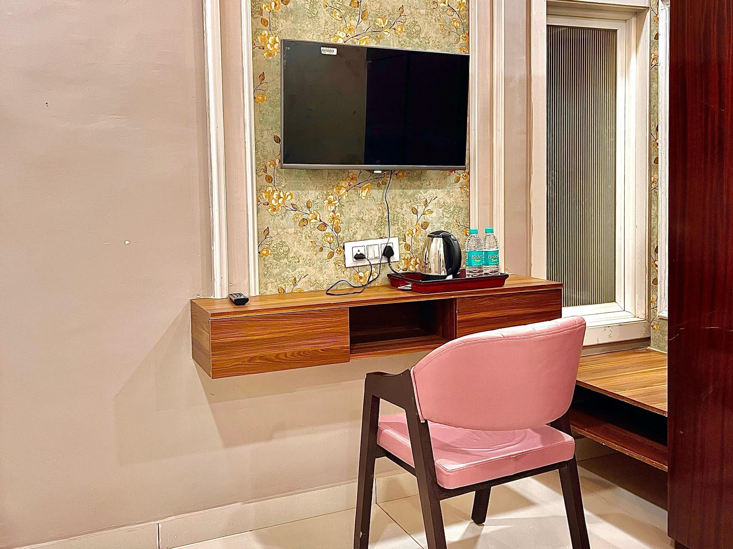 Une pièce avec un bureau en bois, une chaise rose, un téléviseur fixé au mur et une fenêtre avec des stores.