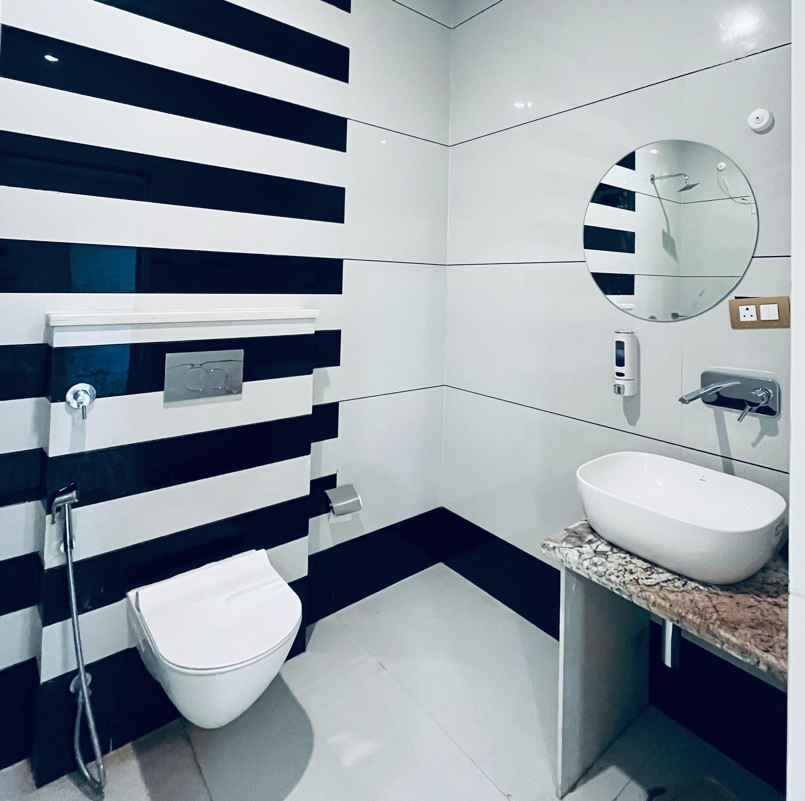 A modern bathroom with a black and white striped wall, a white toilet, a sink with a marble countertop, and a round mirror.