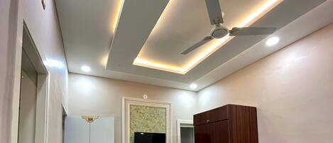 A modern room with a ceiling fan, recessed lighting, a flat-screen TV, and a wooden wardrobe.