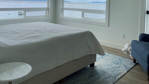 A bedroom with a large bed, a small table, a blue couch, and a view of the sea through large windows. - Luxury 4BR Beachfront Retreat. Designer Kitchen, Views, Game Room, Piano, A/C (Tulalip Bay)