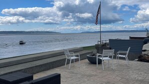 An outdoor seating area with white chairs and a flagpole, overlooking a body of water with a boat and distant land. - Luxury 4BR Beachfront Retreat. Designer Kitchen, Views, Game Room, Piano, A/C (Tulalip Bay)
