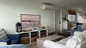 A modern living room with a sofa, a chair, a coffee table, and a bookshelf. There is a painting on the wall and an air conditioning unit mounted on the wall. - Luxury 4BR Beachfront Retreat. Designer Kitchen, Views, Game Room, Piano, A/C (Tulalip Bay)