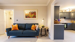 A modern living room with a blue sofa, a small round table with a lamp, and a kitchen area with grey cabinets and a countertop. - No 31 Killarney Holiday Village for 8! Ideal Location! (Killarney)
