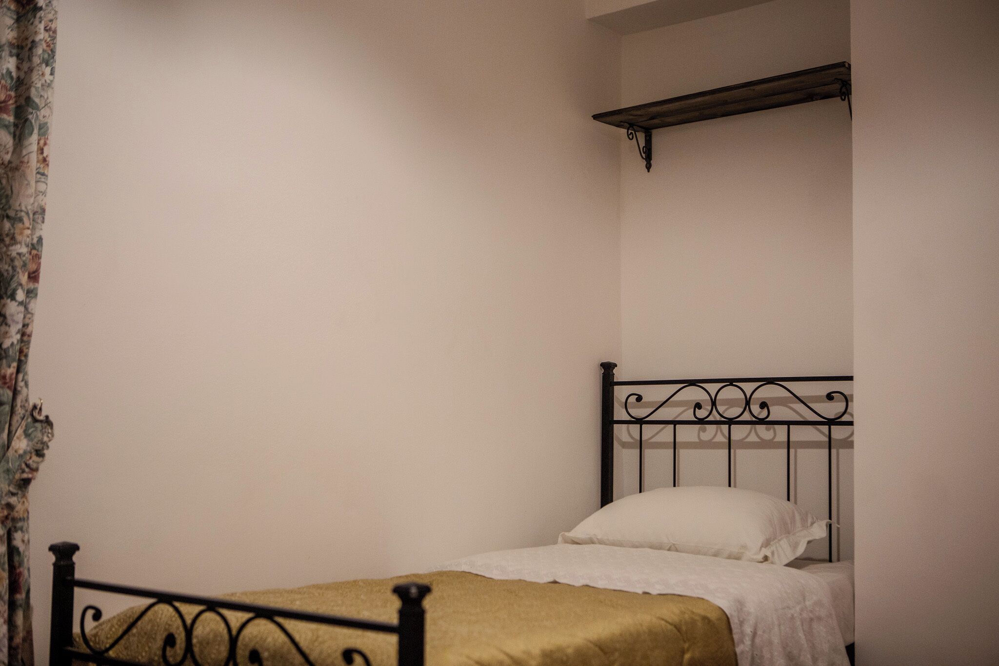 A single bed with a metal frame and white bedding, a wooden shelf above it, and a plain wall.