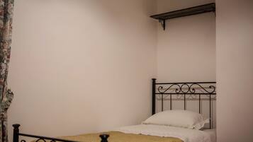 A single bed with a metal frame and white bedding, a wooden shelf above it, and a plain wall.