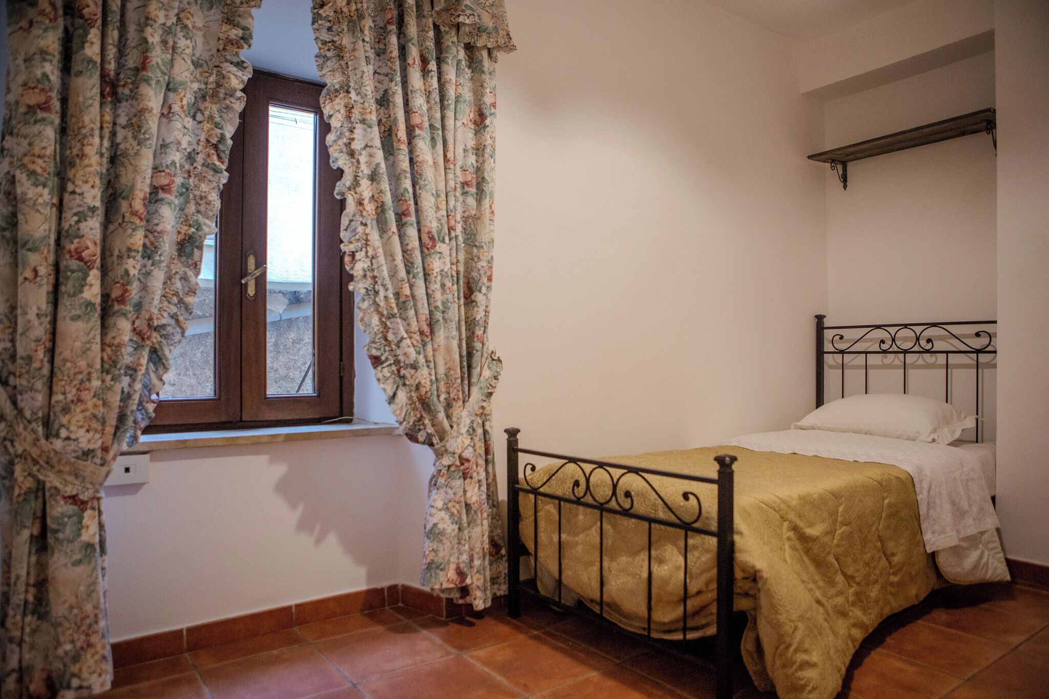 Een slaapkamer met een enkel bed, bloemen gordijnen, een raam en een metalen hoofdbord.