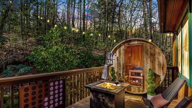 A wooden deck with a fire pit and lounge chairs, surrounded by a forested area.