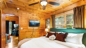 A wooden cabin bedroom with a bed, a ceiling fan, a television, and a window with curtains.