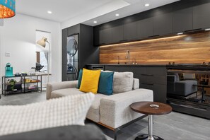 A modern kitchen with dark cabinetry and a wooden backsplash, a grey sofa with colorful cushions, and a small round table. - New Downtown Charlottetown Home Sleeps 5 With 2 Electric Scooters (Charlottetown)