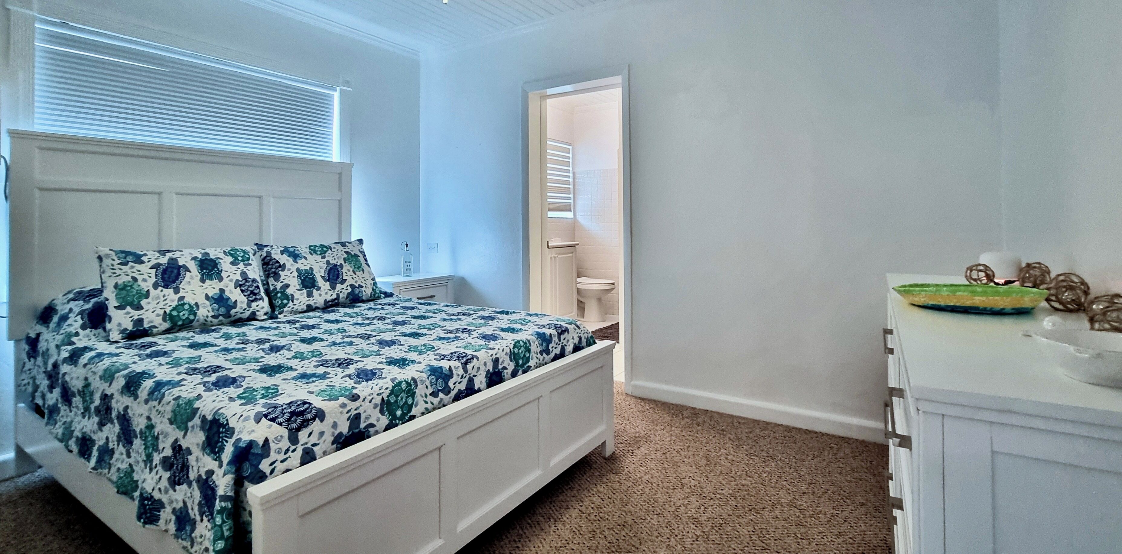 A bedroom with a bed, a dresser, a window, and a door leading to a bathroom.