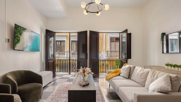 A modern living room with a sofa, armchair, and coffee table. A large TV is mounted on the wall, and a balcony is visible through an open door. - Soho House The Grand Patio (San Juan)