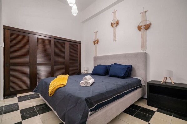A bedroom with a bed, a nightstand, a wall-mounted lamp, and a door with horizontal slats.