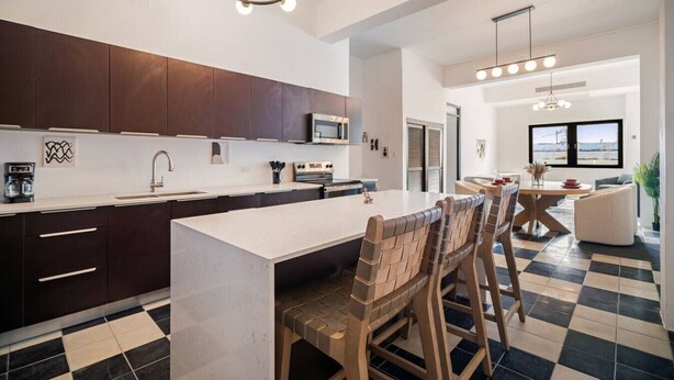 A modern kitchen with dark cabinets, a white countertop, and a central island. There is a dining area with a checkered floor and wooden chairs. - Soho House – Inspired by RH (San Juan)