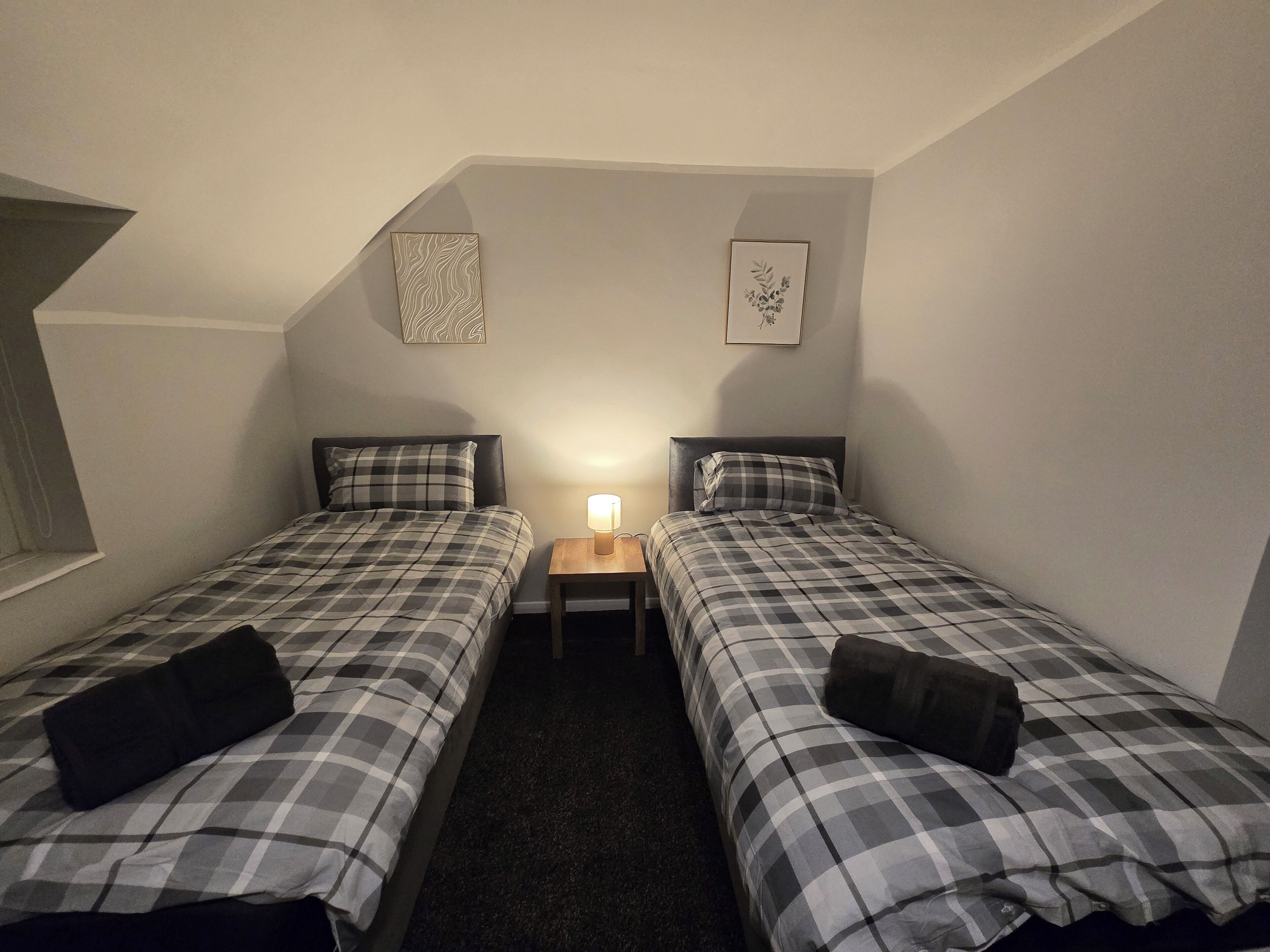 Um quarto pequeno e com pouca luz, com duas camas, cada uma com um cobertor xadrez e um travesseiro. Há uma mesa de cabeceira com uma luminária entre as camas. As paredes são simples e há duas fotos emolduradas penduradas nelas.