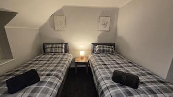Um quarto pequeno e com pouca luz, com duas camas, cada uma com um cobertor xadrez e um travesseiro. Há uma mesa de cabeceira com uma luminária entre as camas. As paredes são simples e há duas fotos emolduradas penduradas nelas.