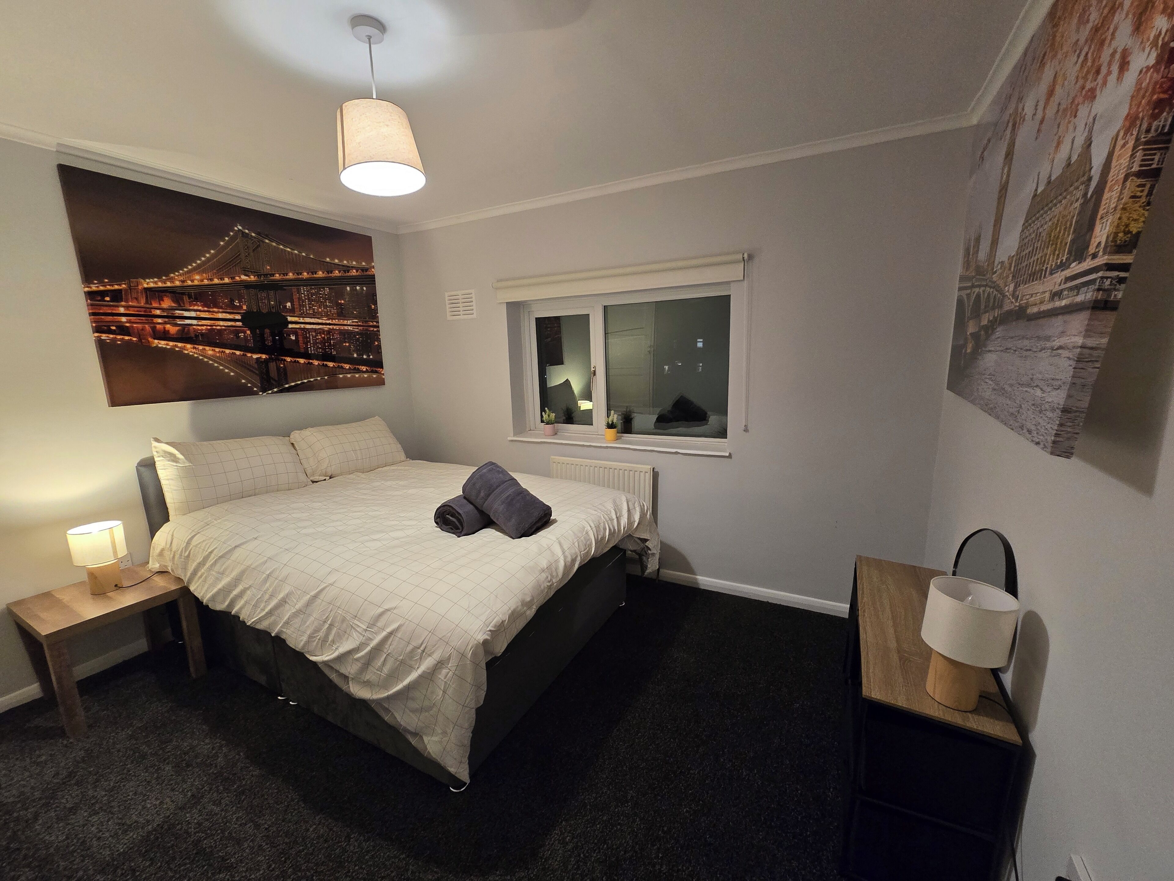 A bedroom with a bed, a nightstand, a lamp, a window with a view of a bridge, and a wall with a cityscape painting.