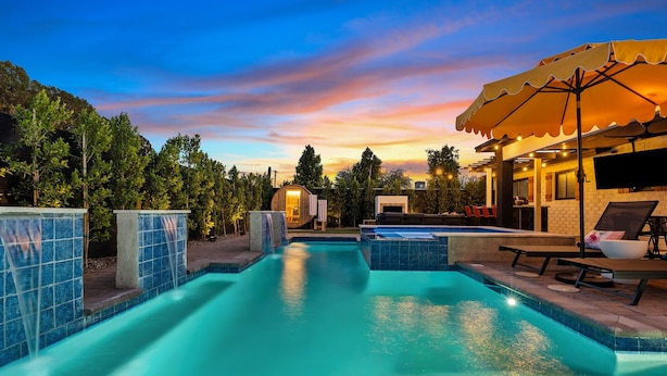 A swimming pool with a view of a sunset, a covered seating area, and a building with a TV screen. - Old Town | Pool | Spa | Sauna | City of Sunsets (Scottsdale)