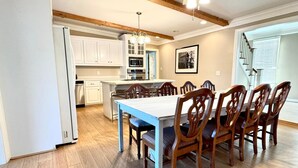 Un comedor amplio con una mesa larga y sillas de madera, una cocina con armarios blancos y una isla central, y una escalera que sube a un nivel superior.