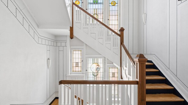 Una escalera con barandillas blancas y escalones de madera, que lleva a un rellano con vistas a una habitación con un gran ventanal.