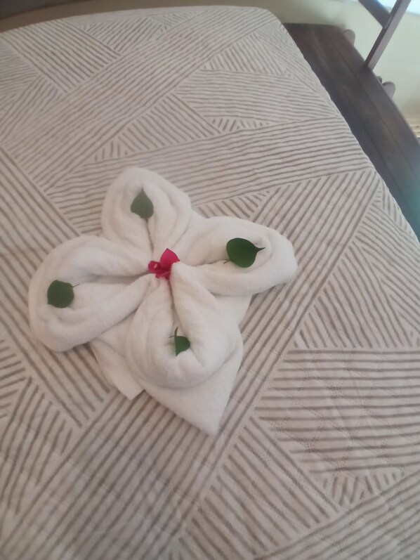 Una cama con un cojín estampado y una toalla dispuesta en forma de flor, adornada con hojas verdes y un lazo rojo.