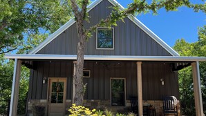 A rustic barn-style building with a porch, stone foundation, and a large tree in front.