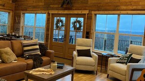 A log cabin interior with a high ceiling, wooden walls, a chandelier, a fireplace, and large windows offering a view of the outdoors.