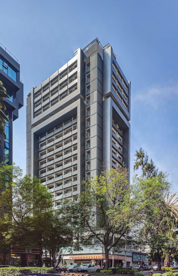 A modern high-rise building with a grid of balconies, surrounded by trees and parked cars.