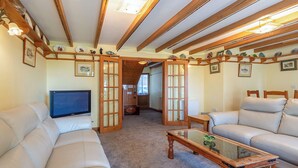 Ein geräumiges Wohnzimmer mit einer großen Ledersofa, einem Glastisch und einem Fernseher. Das Zimmer verfügt über Holzbalken an der Decke und einen Blick in einen anderen Raum durch eine Glastür.