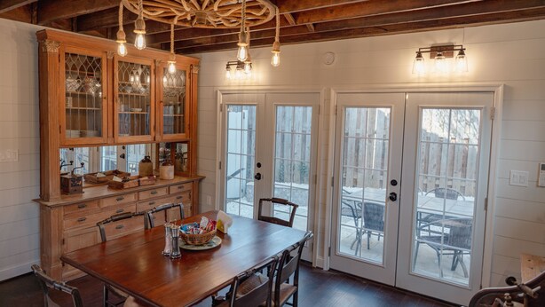 A dining area with a wooden table, chairs, a large cabinet with glass doors, and a ceiling with exposed beams and hanging lights.