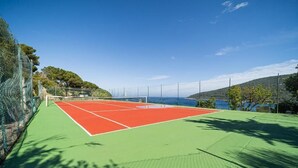 A tennis court with a red playing surface and green surrounding area, surrounded by a fence and trees. - Villa with private beach and pool (Toscana)