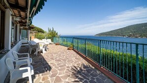 A terrace with stone flooring, white plastic chairs, and a view of the sea. - Villa with private beach and pool (Toscana)