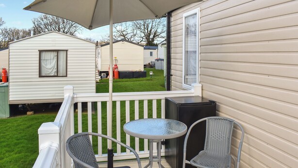Een balkon met twee stoelen en een tafel, een witte leuning en uitzicht op aangrenzende stacaravans.
