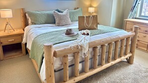 A wooden bed with a headboard, two bedside tables, a window, and a nightstand with a lamp. - Modern Rustic 3 Bedroom with Pool and Hot tub (Hailey)