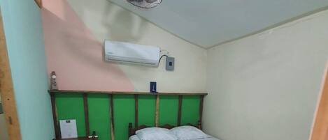 A small, single-bed room with a green wall, a wooden headboard, a bedside table, an air conditioner, and a ceiling fan.