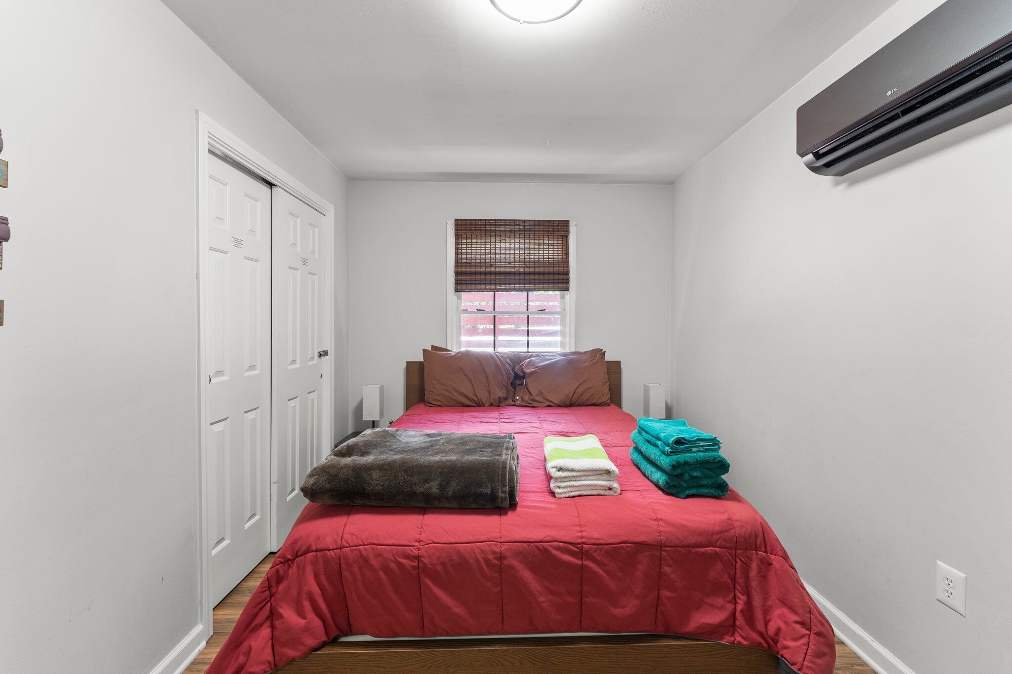 Un dormitorio con una colcha roja, una ventana con persianas, un calentador y una puerta con cerradura.