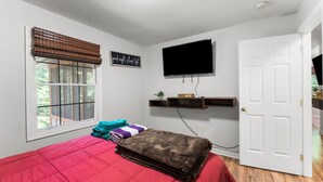 A bedroom with a red bedspread, a window with blinds, a mounted TV, and a door.