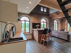Eine Küche mit Holztischplatte, Spüle und einer Bar mit Hockern. Es gibt Rundbogenfenster und eine Steinwand.